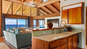 A spacious living area with a large window overlooking the ocean, a wooden ceiling, and a kitchen with a wooden island. - Summer Deal Kona Oceanfront Estate with Pool Water Slide Guest House (Kauilua Kona)