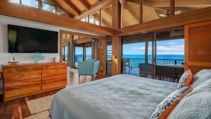 A spacious bedroom with a large bed, wooden ceiling beams, a television, and a view of the ocean. - Summer Deal Kona Oceanfront Estate with Pool Water Slide Guest House (Kauilua Kona)