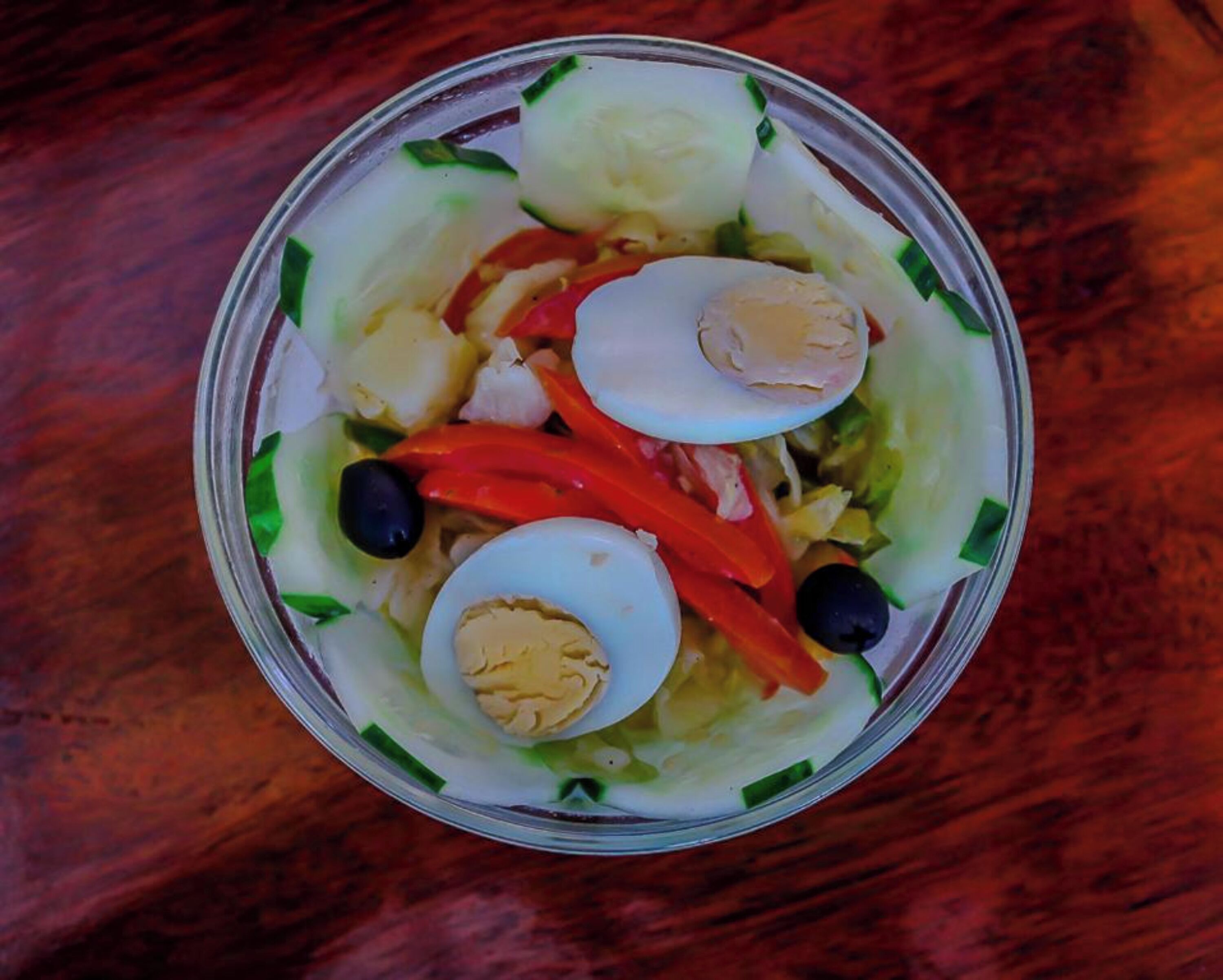 A salad with cucumbers, red peppers, black olives, and hard-boiled eggs in a clear glass bowl.