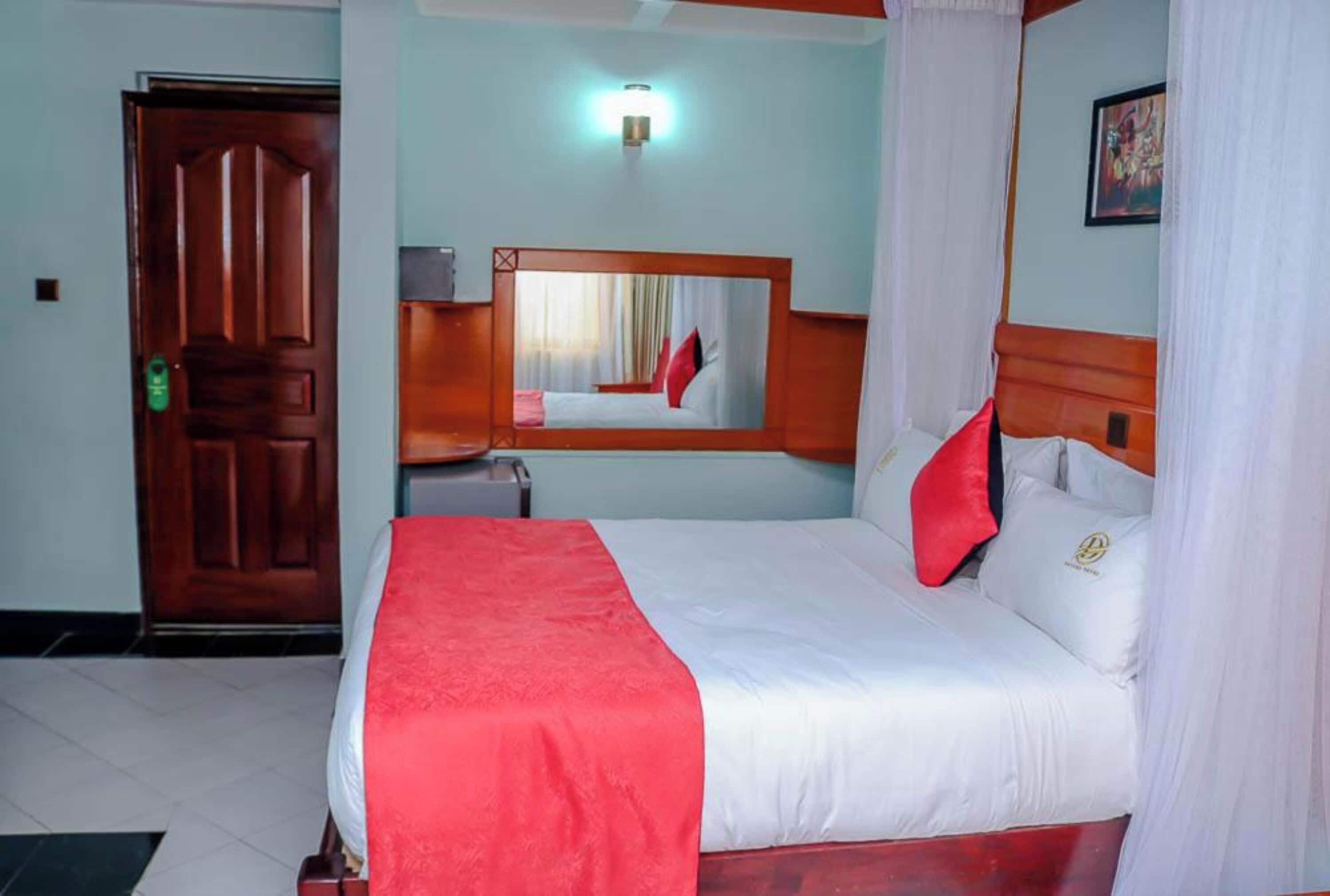 Habitación de hotel con una cama grande, una puerta de madera, una mesita de noche, un espejo y una lámpara fijada a la pared.