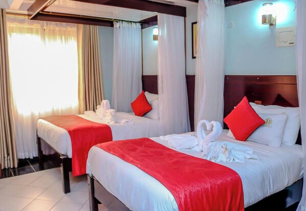 A hotel room with two beds, each with white bedding and red pillows, and a red blanket. The room has a wooden headboard and a large window with curtains.