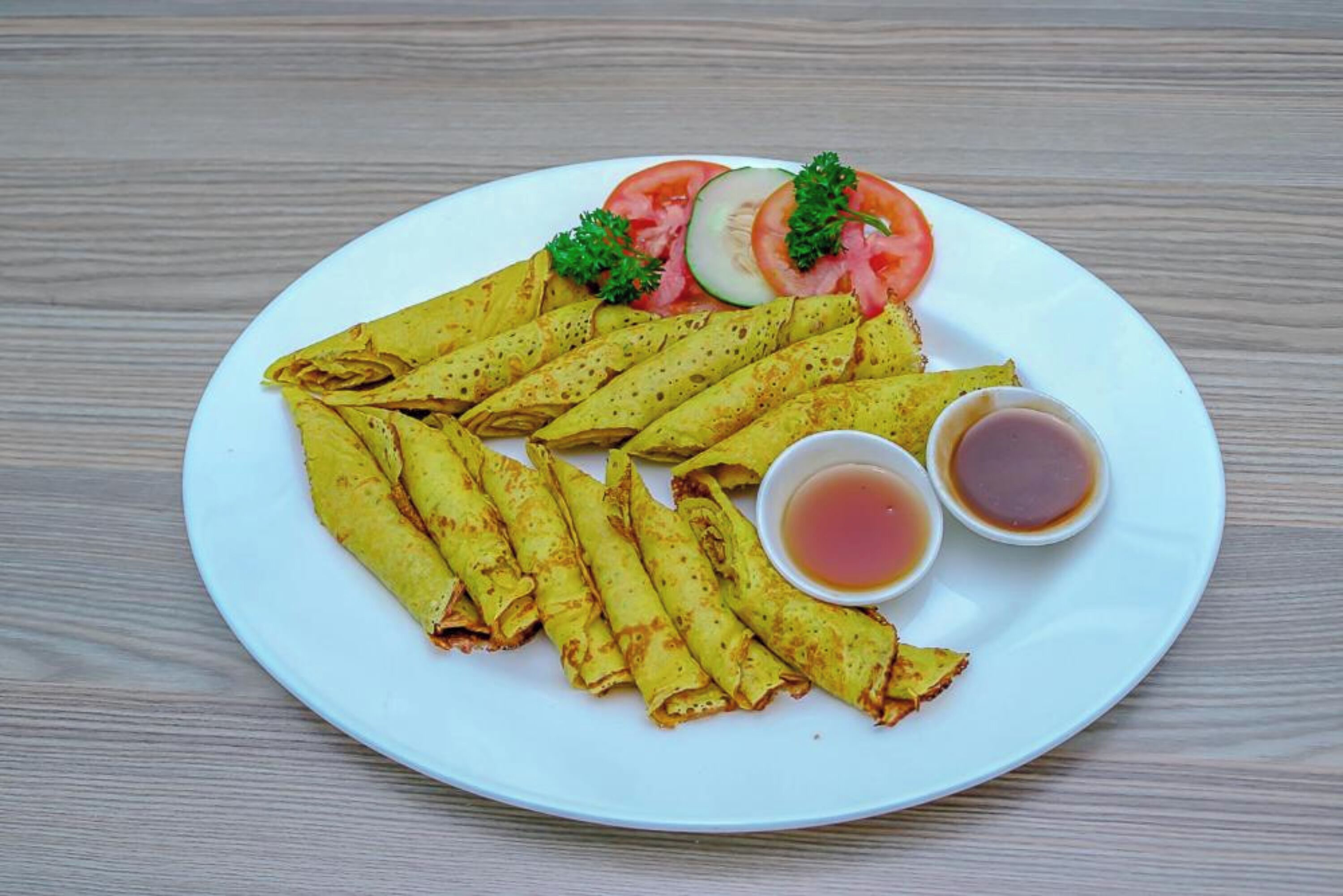 Un plato de crepas enrolladas con rodajas de tomate, pepino y perejil, acompañado de dos tazones pequeños de jarabe.