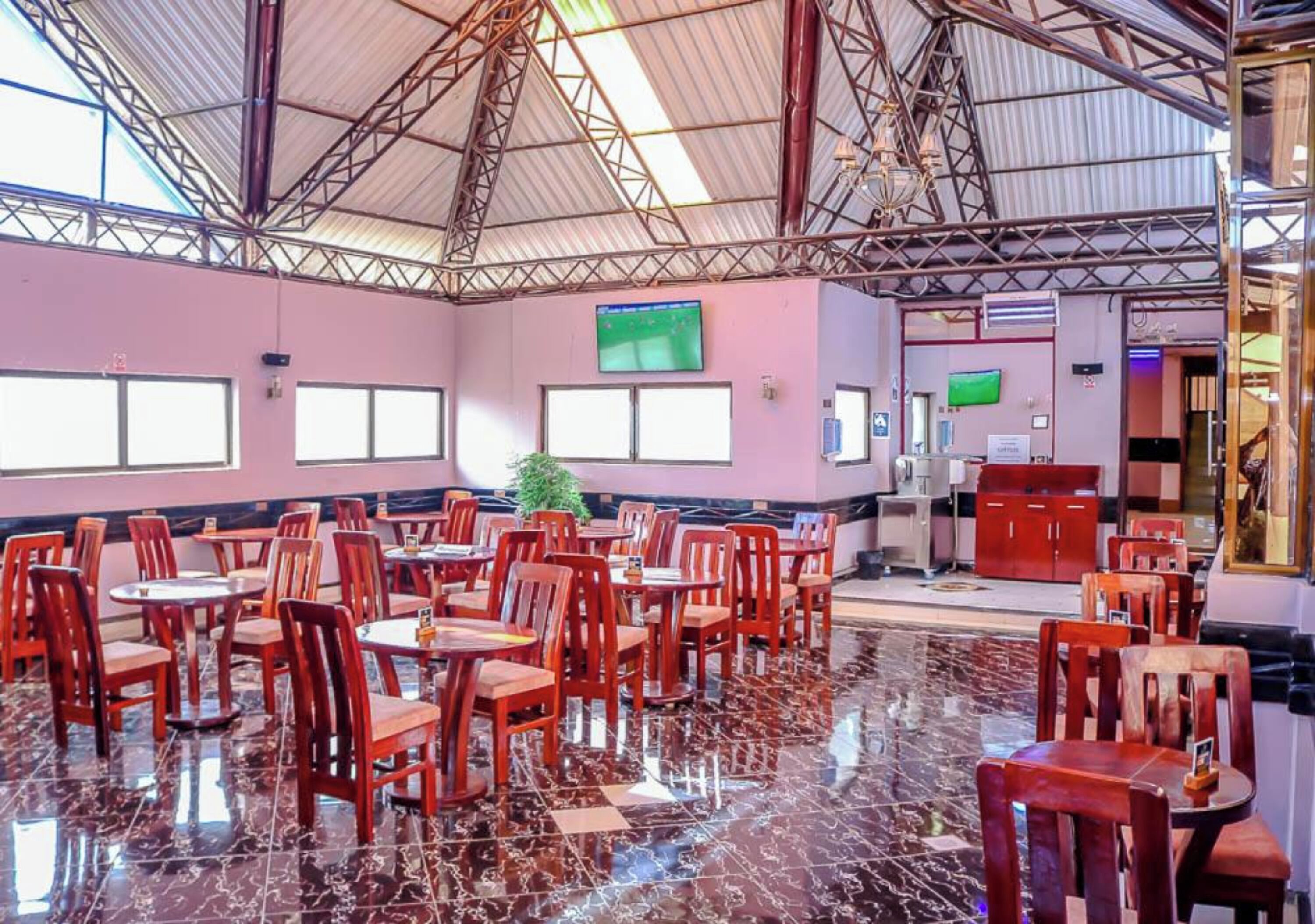 A spacious indoor area with multiple tables and chairs, a high ceiling with metal beams, and a large screen on the wall.
