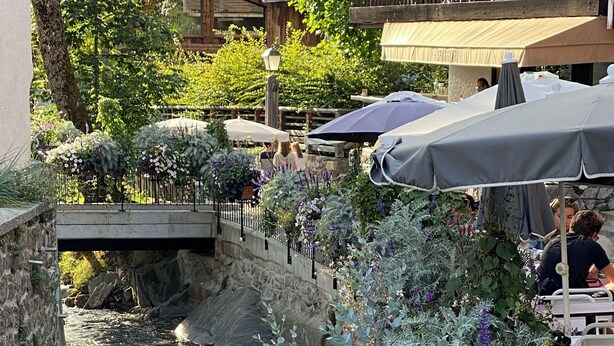 Ein Restaurant am Flussufer mit Außenbestuhlung, einer Stegbrücke und üppiger Vegetation.