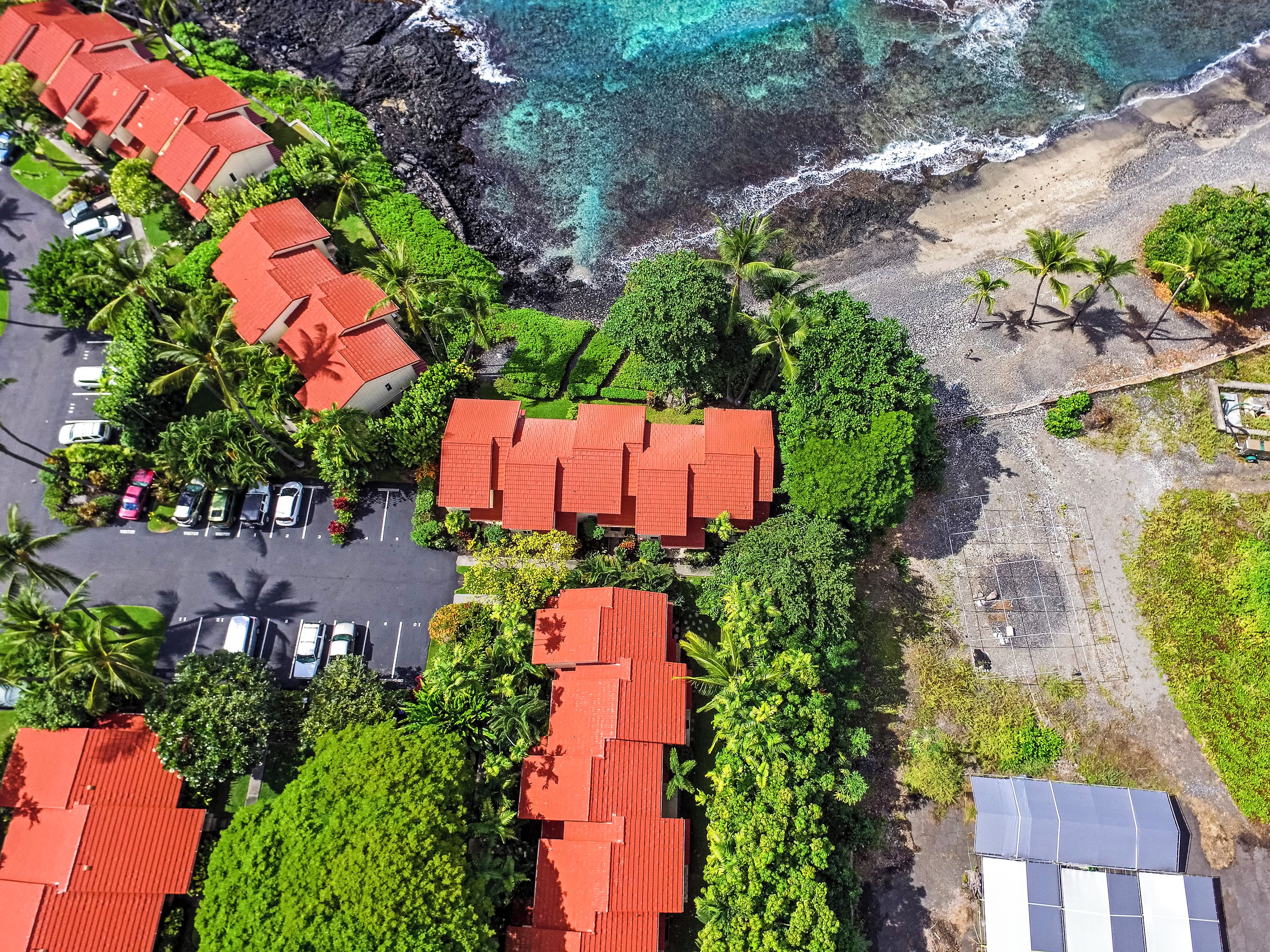 An aerial view of a coastal area with multiple red-roofed buildings, a parking lot, and a beach with palm trees.