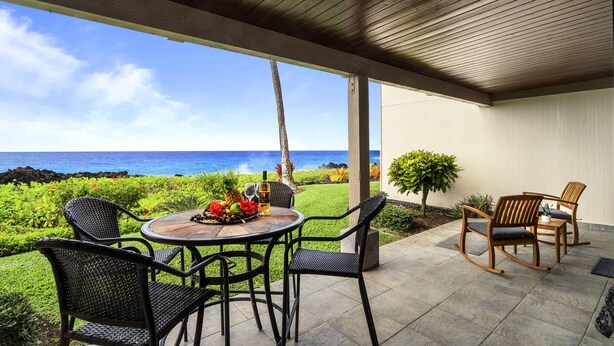 A covered patio with a round table and chairs, overlooking a lush garden and the ocean.