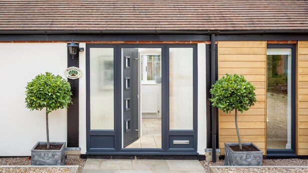 A modern building with a dark-colored door, two potted trees, and a sign on the wall. - Cotswold Barn Conversion With Private Hot Tub (Pershore)