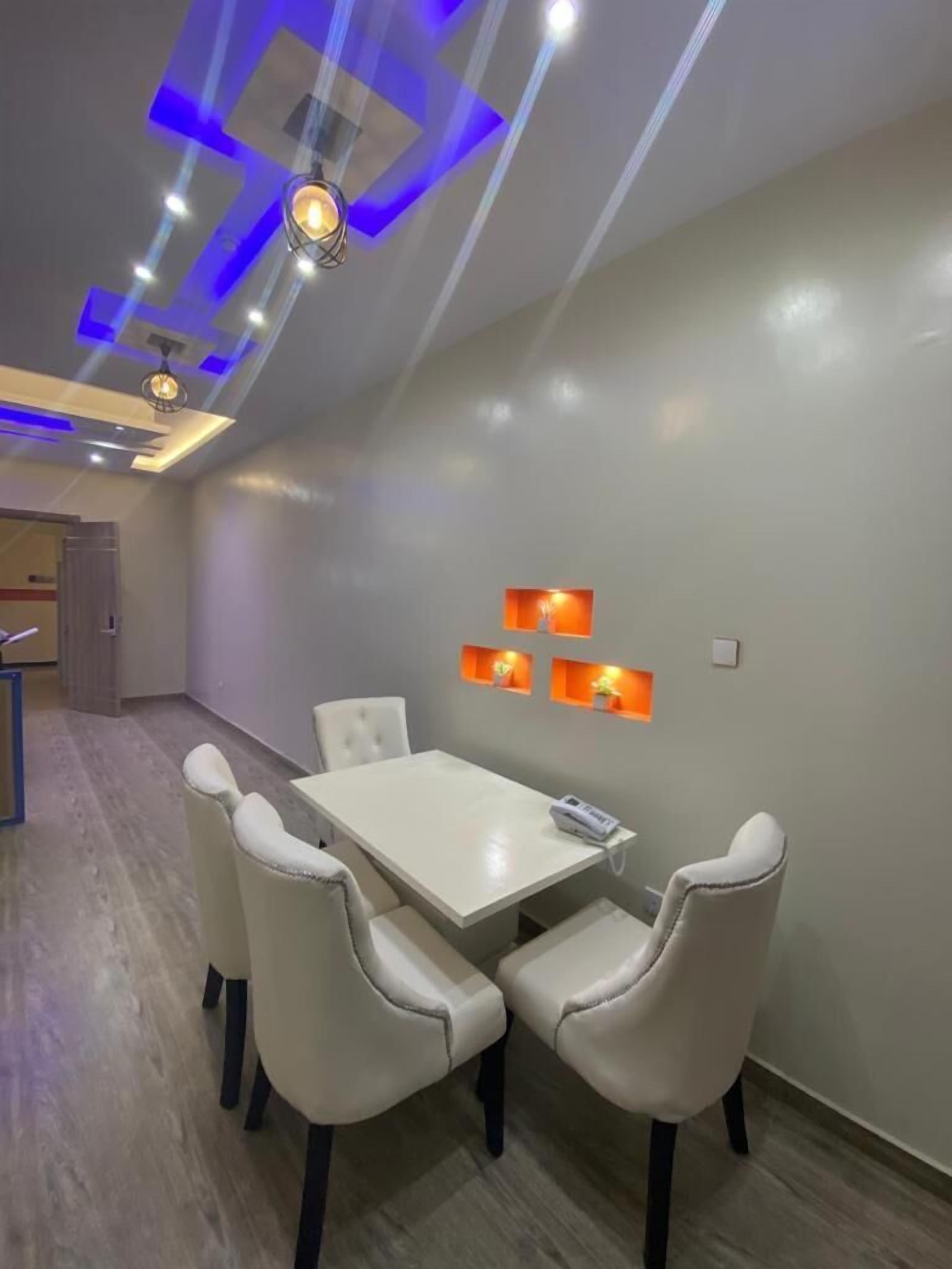 A dining area with a white table and four chairs, a ceiling with blue lighting, and wall-mounted orange lights.