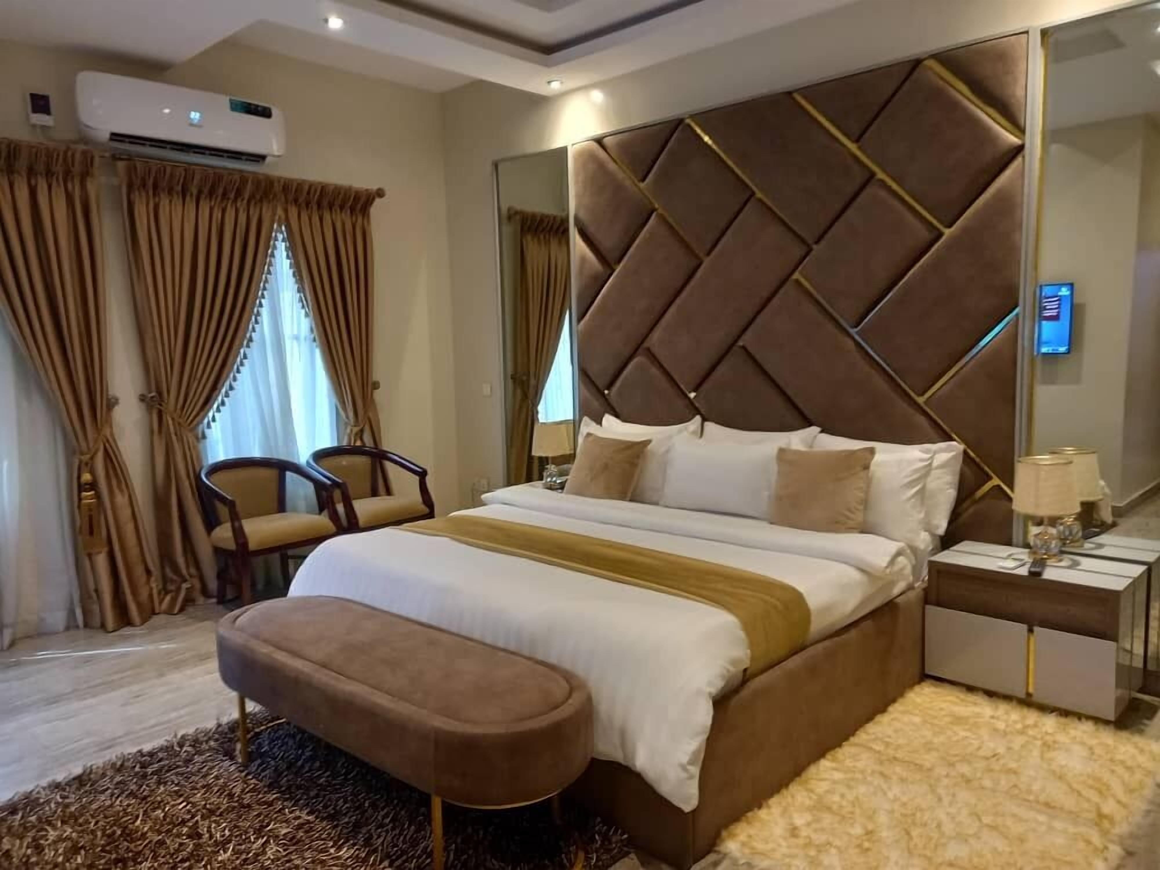 A modern hotel room with a large bed, a headboard with a geometric pattern, a bedside table with lamps, and a large mirror.