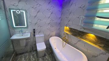 Ein Badezimmer mit Wand in Marmoroptik, einem weißen Waschbecken, einer Toilette und einer freistehenden Badewanne mit goldfarbenen Armaturen.
