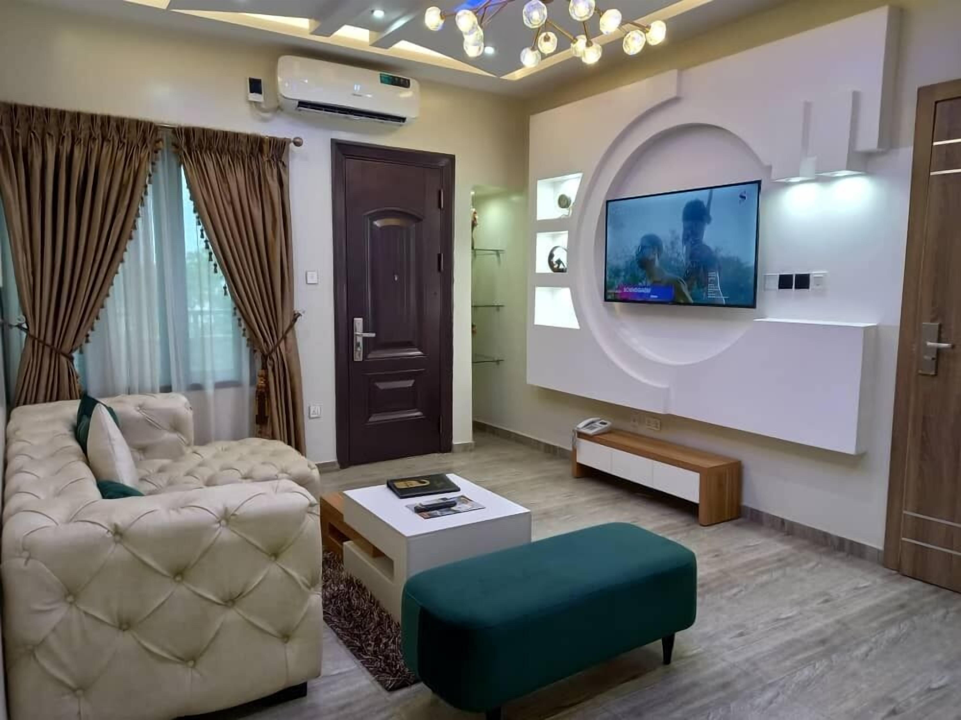 A modern living room with a beige sofa, a teal ottoman, a white coffee table, and a mounted TV.