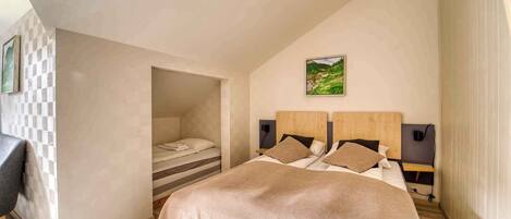 Quarto com teto inclinado, uma cama com manta bege, duas mesas de cabeceira com luminárias e um quadro na parede.