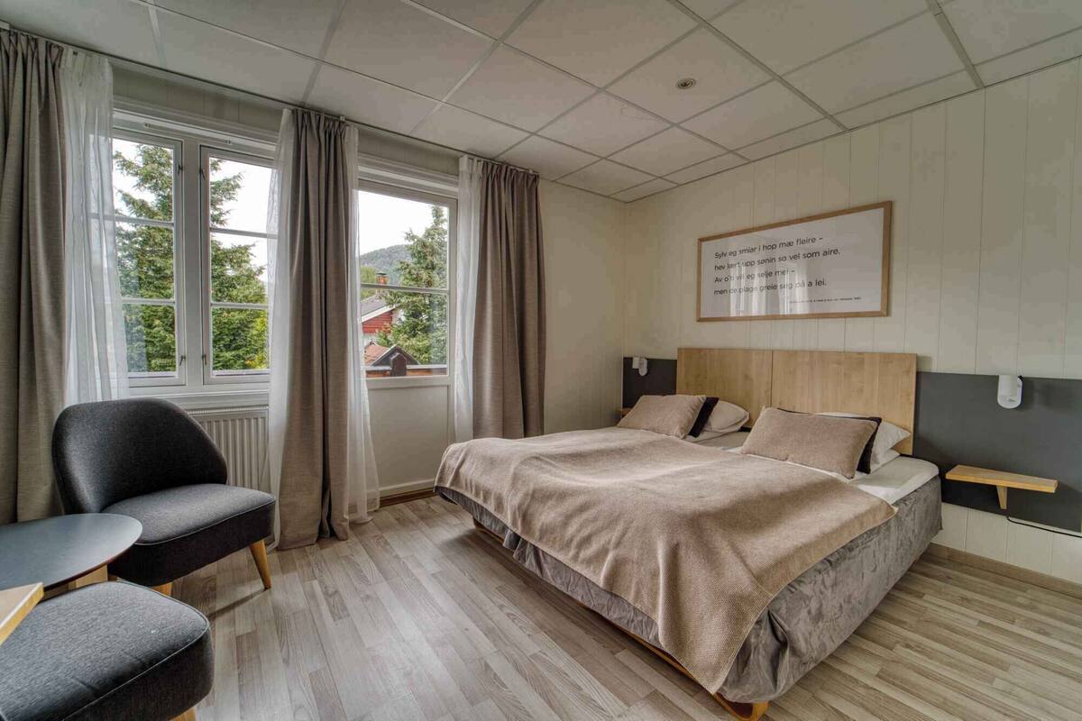 A bedroom with a bed, a chair, a window with curtains, and a wall-mounted light fixture.