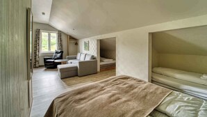 A spacious loft bedroom with a large bed, a sofa, and a chair. There is a window with curtains, a radiator, and a small table.