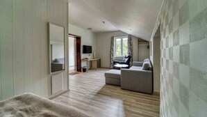 A spacious room with a high ceiling, a large window with curtains, a flat-screen TV mounted on the wall, and a seating area with a sofa and armchairs.