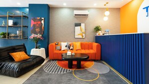 Un moderno lobby de hotel con un sofá de cuero negro, un sofá naranja y una mesa de centro negra. Hay una mesa redonda blanca con un jarrón de flores y una planta pequeña. Las paredes están pintadas de azul y naranja, y hay dos cuadros colgados.