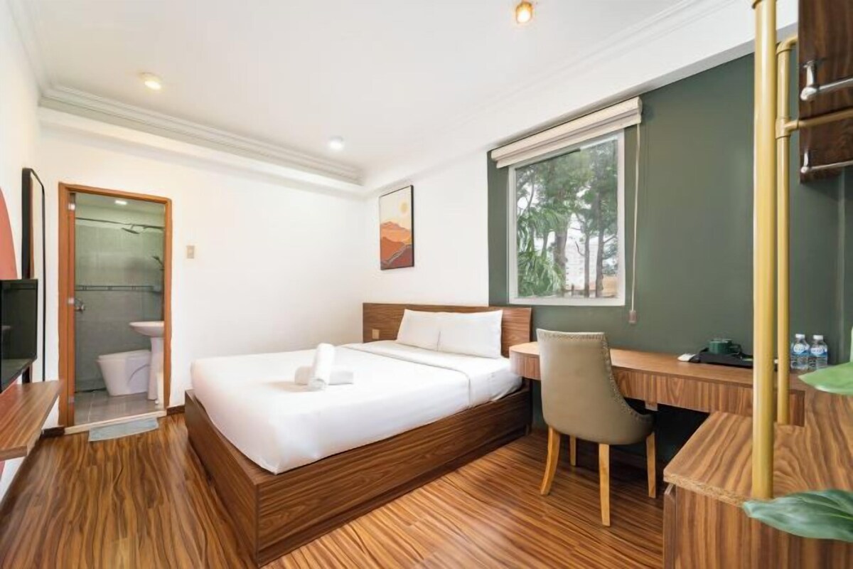 A hotel room with a bed, a desk, a chair, a window, and a door leading to a bathroom.