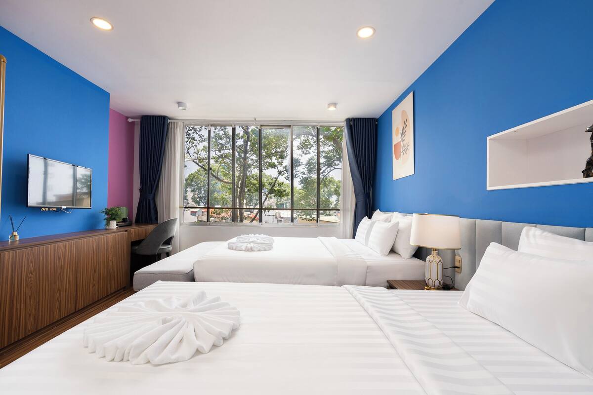 A hotel room with two beds, a TV, a desk, and a window with a view of trees.