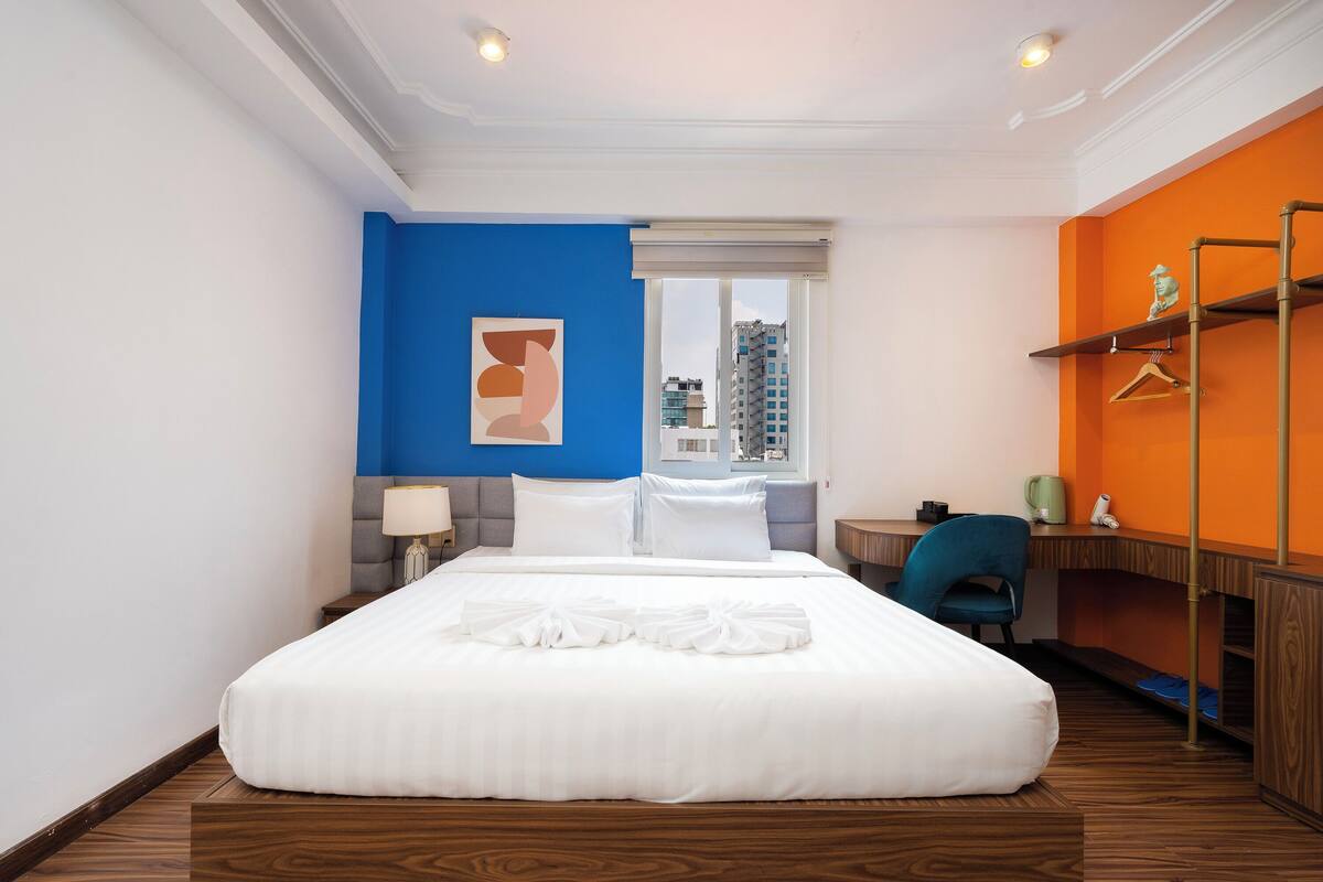 A hotel room with a large bed, a desk, a chair, a window, and a view of buildings outside.