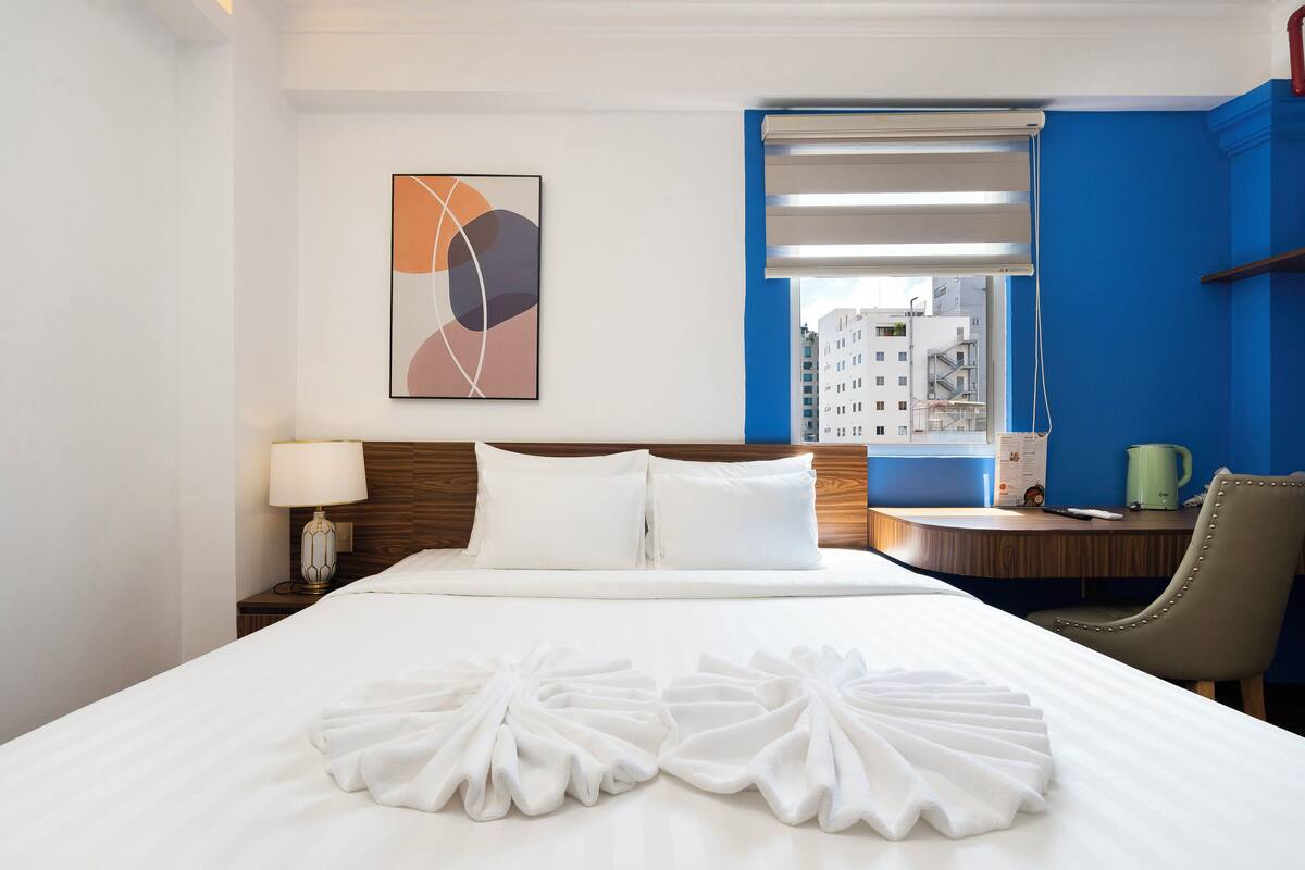 A hotel room with a neatly made bed, a desk with a chair, a lamp, and a window with blinds.