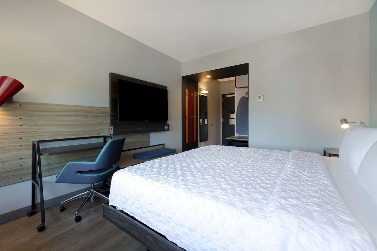 Ein modernes Hotelzimmer mit einem großen Bett, einem Schreibtisch mit Stuhl, einem flachbildfernseher an der Wand und einem begehbaren Kleiderschrank.