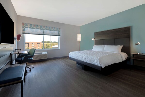 A hotel room with a large bed, a desk with a chair, a TV, and a window with a view of buildings.