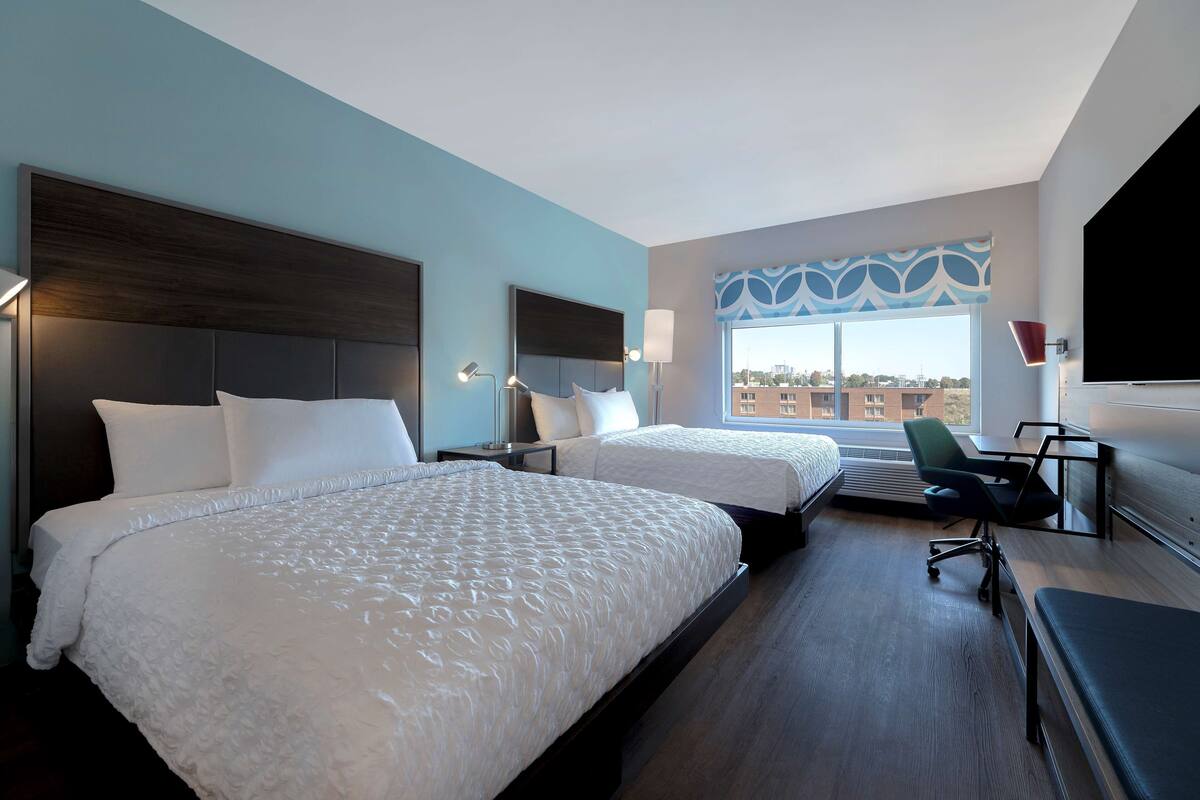 Ein Hotelzimmer mit zwei Betten, einem Schreibtisch, einem Sessel und einem großen Fenster mit Blick auf die Stadt.