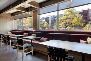 Een modern café met houten tafels en bankjes, grote ramen met uitzicht op gebouwen en bomen.