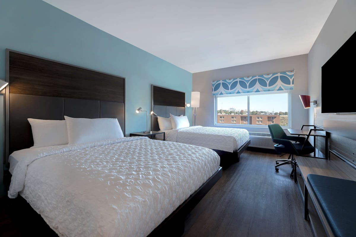 Ein Hotelzimmer mit zwei Betten, einem Schreibtisch, einem Sessel und einem großen Fenster mit Blick auf die Stadt.