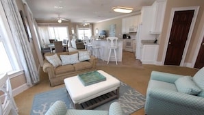 Amplia sala de estar con un sofá, reposapiés y dos sillones. Se ve una cocina y comedor al fondo.