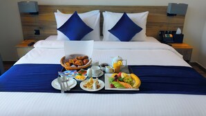 A neatly made bed with a blue bedspread, two pillows, a bedside table with a lamp, and a tray with breakfast items including croissants, fruit, and coffee. - Kingsgate Canal Hotel (Dubai)
