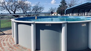Piscina rectangular elevada con un cartel de seguridad, rodeada de un patio de ladrillo y árboles.