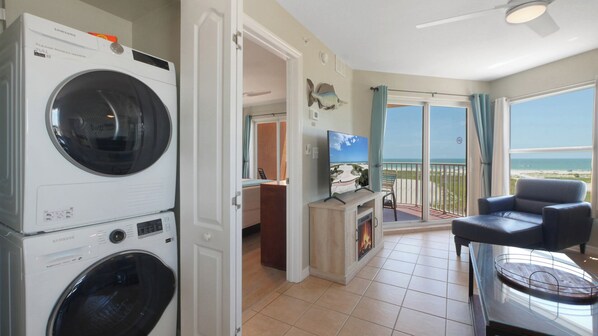A room with a washing machine, a television, a balcony with a view of the beach, and a chair.
