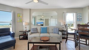 A living room with a sofa, a coffee table, a blue armchair, a dining table with chairs, and a view of the ocean through large windows. - Surf Beach 506 - Spectacular Sunsets - Beach Front (Treasure Island)