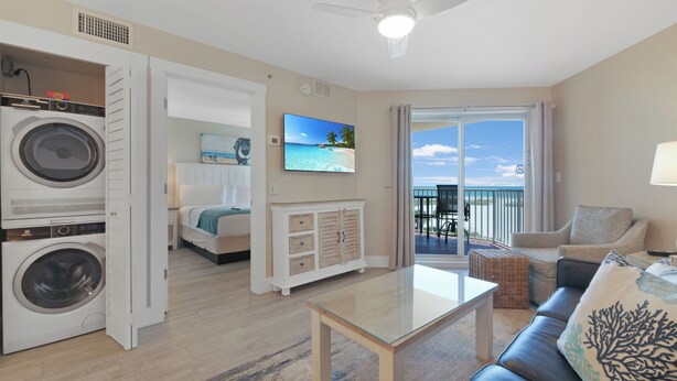 Habitación de hotel con balcón, cama, lavadora, mesa de centro de vidrio y vistas al océano.