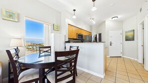 A compact kitchen with wooden cabinets, a microwave, and a refrigerator. There is a dining area with a glass table and chairs, and a view of the ocean through a large window. - Surf Beach 606 - Spectacular Sunsets - Beach Front (Treasure Island)