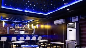 A bar area with a brick wall, a white refrigerator, and a 'No Smoking' sign. There are several round tables and chairs arranged for seating. - Pasadena Suites (Lekki)