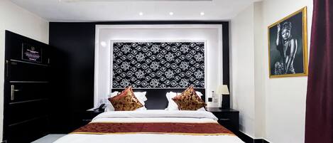A modern hotel room with a large bed, black and white patterned headboard, and a red bed skirt. There is a nightstand with a lamp and a framed picture on the wall.