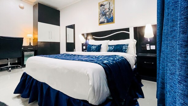A hotel room with a large bed, a desk, a chair, a TV, and a painting on the wall. - Pasadena Suites (Lekki)