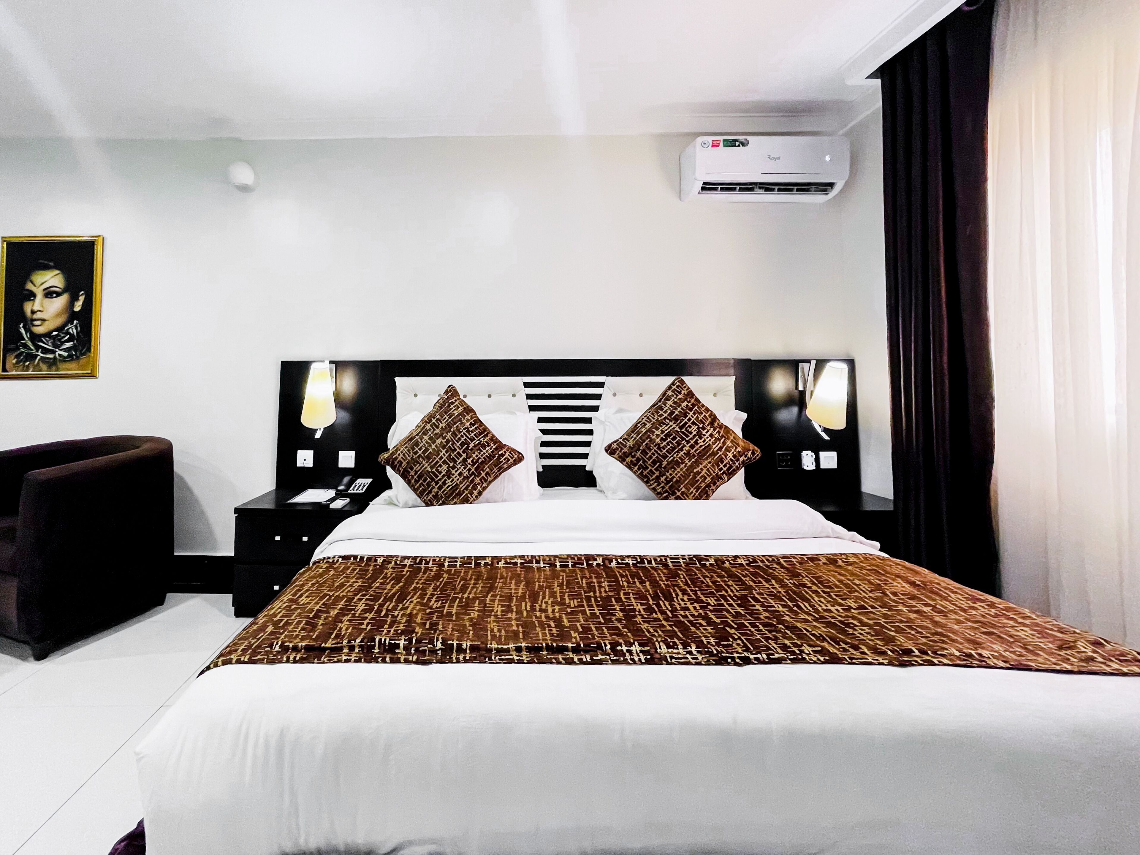 A hotel room with a large bed, a bedside table, a chair, an air conditioning unit, and a painting on the wall.