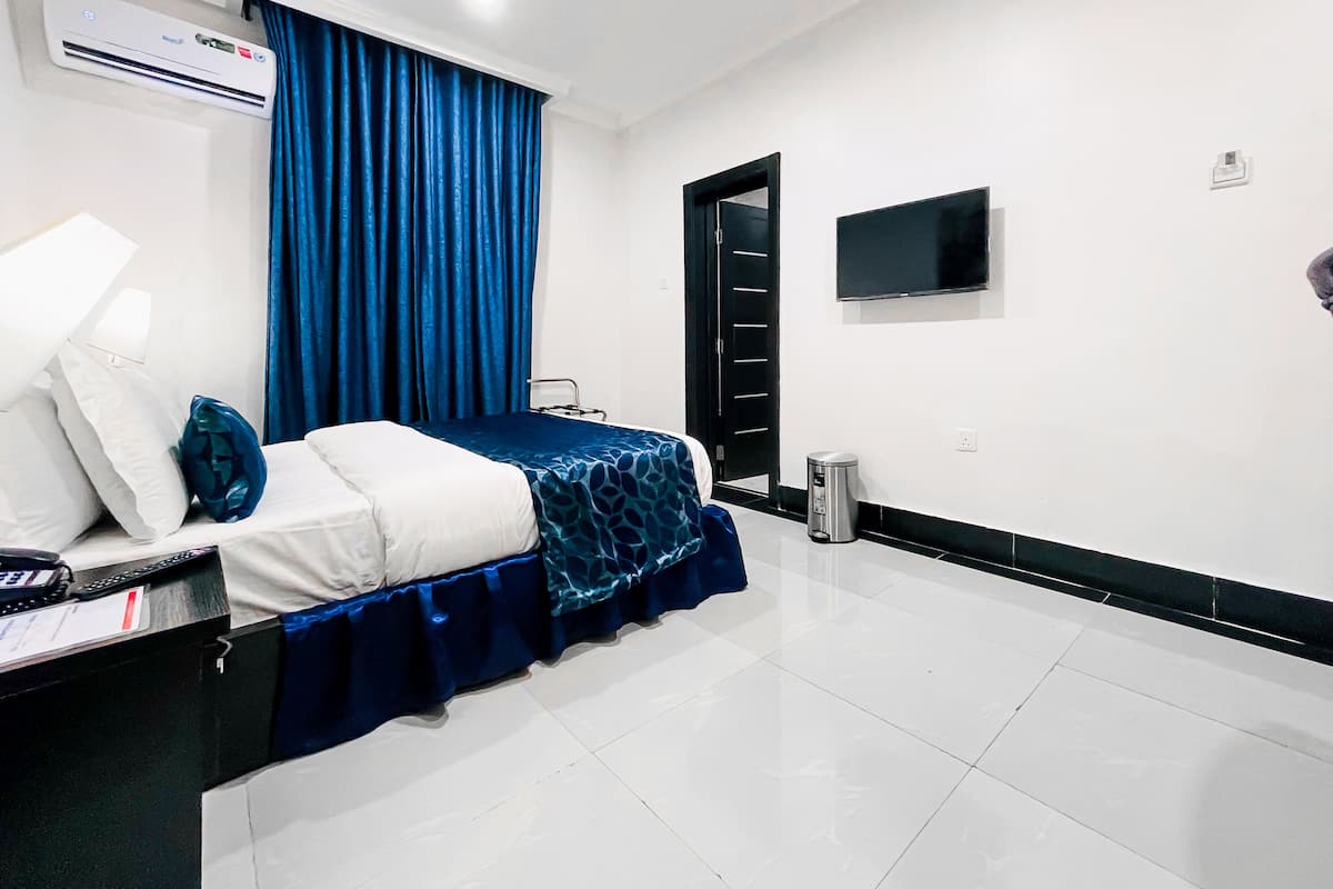 A hotel room with a bed, a bedside table, a lamp, a TV mounted on the wall, and a trash bin.