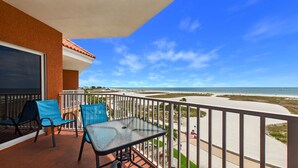 A balcony with a glass table and chairs, overlooking a beach with a clear sky.