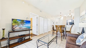 A living room with a TV, a glass coffee table, a sofa, and a dining area with chairs.