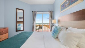 A hotel room with a bed, a dresser, a balcony with a view of the beach, and a framed picture on the wall.