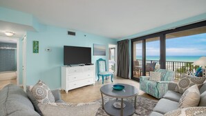 Ein geräumiges Wohnzimmer mit einem großen Flachbildfernseher, einer weißen Kommode, einem blauen Sessel und einem Balkon mit Blick auf den Strand.