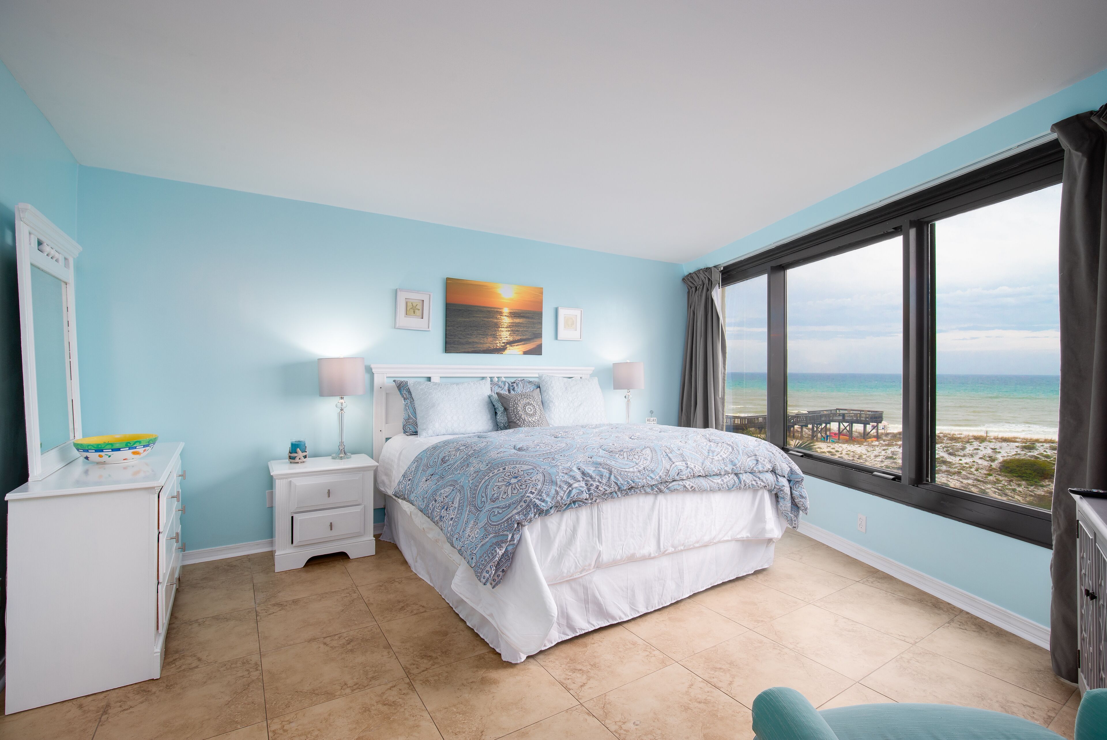 A bedroom with a bed, nightstands, a dresser, and a view of the beach through large windows.