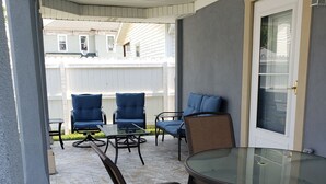 A covered patio area with a glass table and chairs, a white door with a glass panel, and a white fence in the background. - La Casita Guest House (Muscatine)