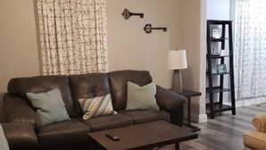 A living room with a brown leather sofa, a coffee table, and a lamp. There is a bookshelf with a 'Wi-Fi' sign and a picture frame. - La Casita Guest House (Muscatine)