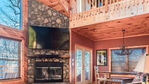 A spacious living area with a stone fireplace, wooden floors, a large flat-screen TV, and a balcony with a railing. - Comfort & Relaxation 3 br Cabin Wraparound Decks Amazing Views 2 Hot Tubs (Pigeon Forge)