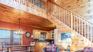 A spacious wooden interior with a dining area, two leather sofas, a staircase, and a kitchen in the background. - Comfort & Relaxation 3 br Cabin Wraparound Decks Amazing Views 2 Hot Tubs (Pigeon Forge)